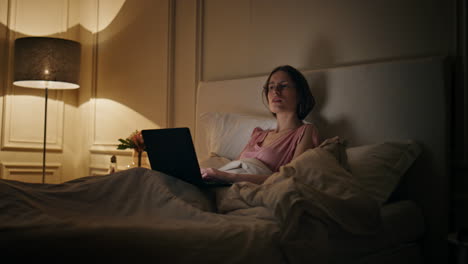late freelancer working home resting in bed. serious woman typing laptop message