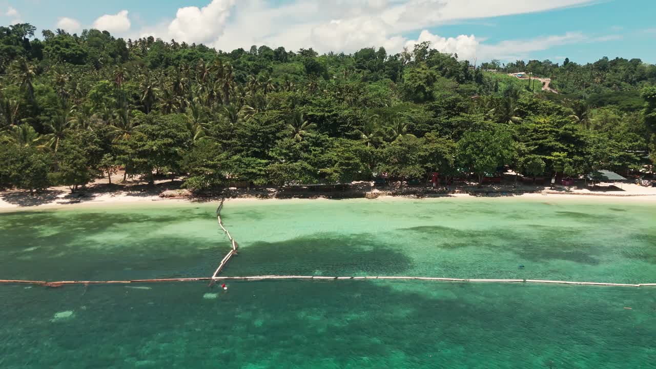 Wide, low-angle aerial shot of an idyllic tropical beach. Clear turquoise and deep blue ocean water transitions to white sand, backed by a dense palm forest and hillside resort buildings
