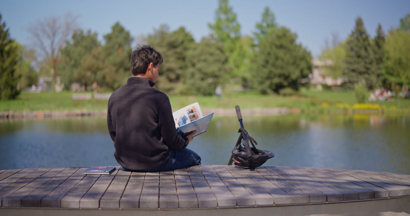 Rearview of young Teenager reading magazine or book near calm lake in public park outdoors