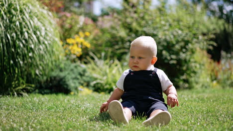 beautiful blond baby boy sits on the grass in the sun. cute child looks around with interest. blurred backdrop.