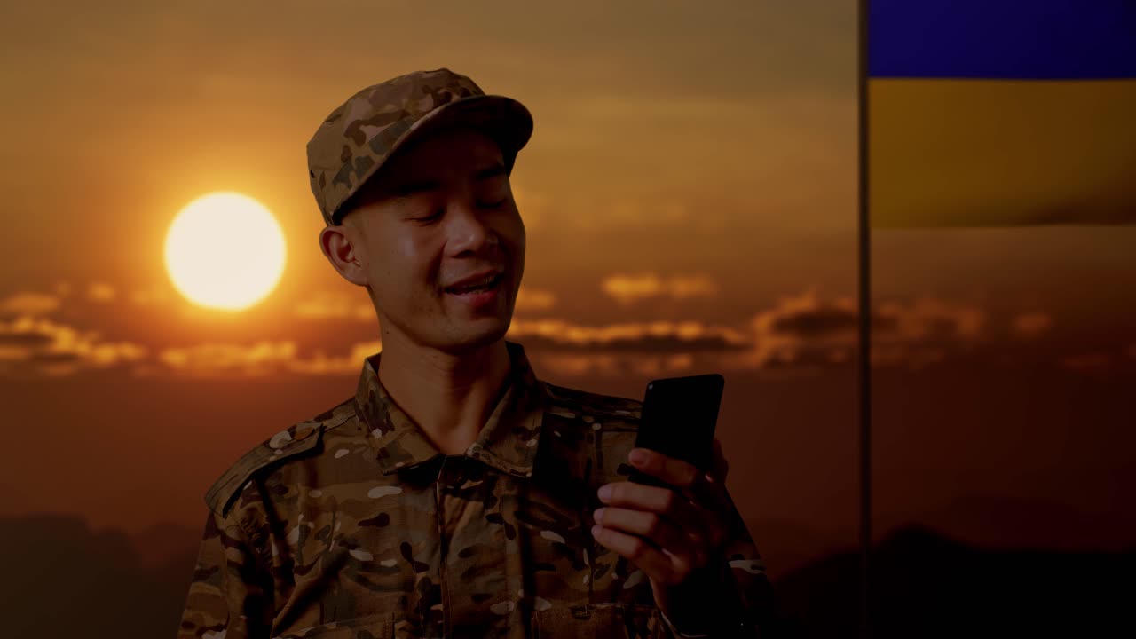 soldado comunicándose a través de un teléfono inteligente durante la puesta del sol con la bandera ucraniana en el fondo