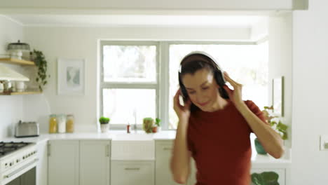 young woman dancing joyfully in kitchen while listening to music on headphones, at home