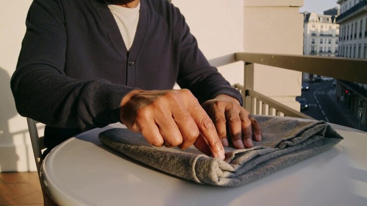 Smoothing man in cardigan after hands rest, tracing folded gray garment on balcony table, preparing