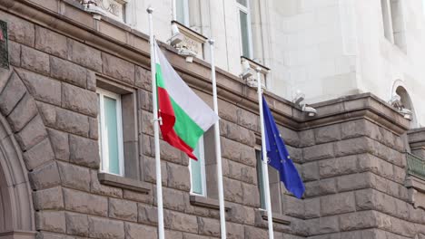 Low-angle-shot-of-an-ancient-government-building-in-Sofia,-Bulgaria-with-Bulgaria-flag-and-EU-flag-visible-in-the-foreground-at-daytime