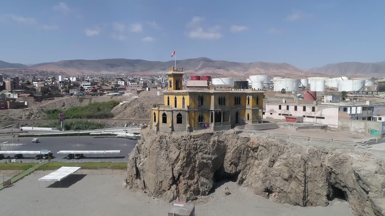 A hauntingly beautiful abandoned mansion sits atop a rugged cliff, contrasting with the urban and industrial backdrop. Ideal for storytelling, travel, and history-themed content. Mollendo, Arequipa.
