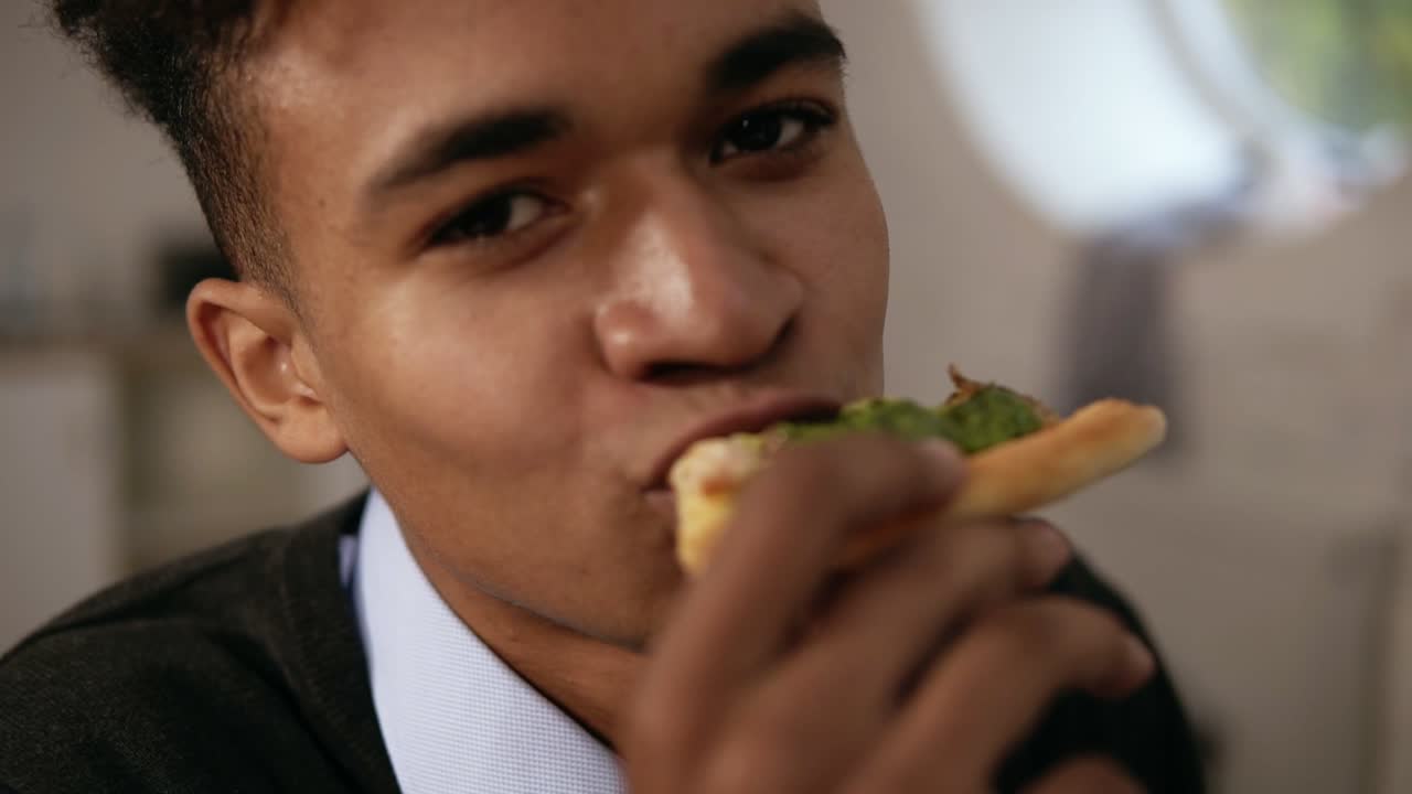 Close Up view of young african american enjoying pizza during lunch at work. He is biting a slice and looking in the camera in