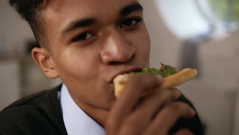 close up view of young african american enjoying pizza during lunch at work. he is biting a slice and looking in the camera in