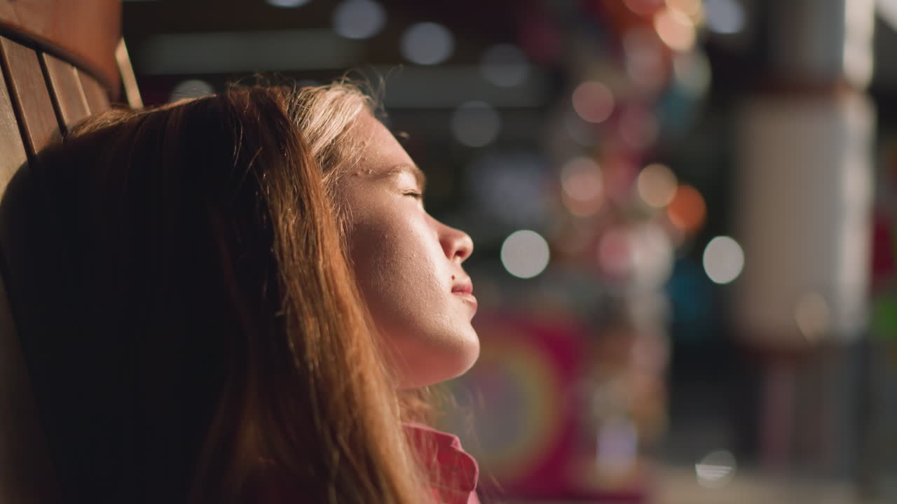 una dama blanca con un vestido rosa se relaja en una silla de madera, la cara muestra espinillas ligeras, mientras que las luces borrosas crean un efecto brillante en el fondo, las personas son visibles pasando en el fondo borroso
