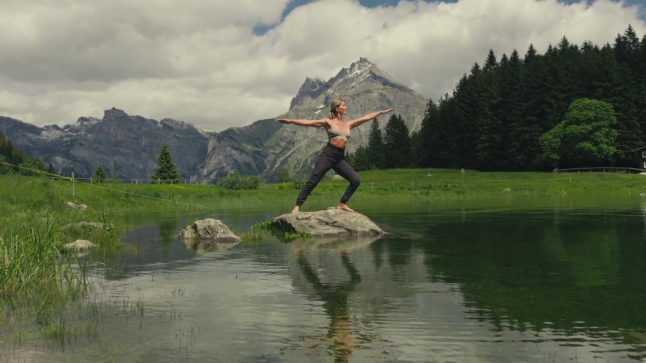 Una donna bionda caucasica esegue una posa yoga del guerriero su una roccia in un lago alpino incontaminato, di fronte alla telecamera. Le spettacolari Alpi Svizzere con cime innevate creano una scena di benessere mozzafiato