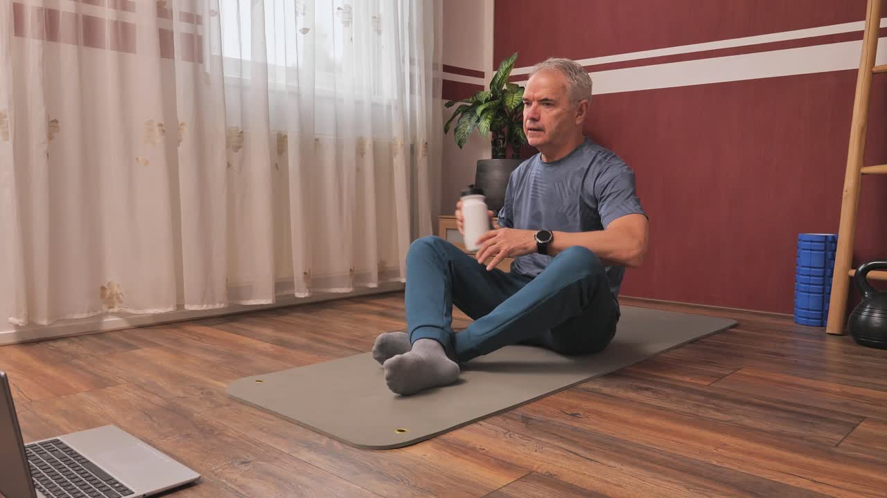 Senior man hydrating after home workout, sitting on mat with laptop and fitness gear nearby