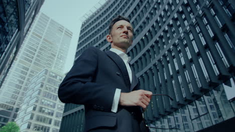 elegante hombre caucásico de pie al aire libre en traje y gafas en la mano, ángulo bajo