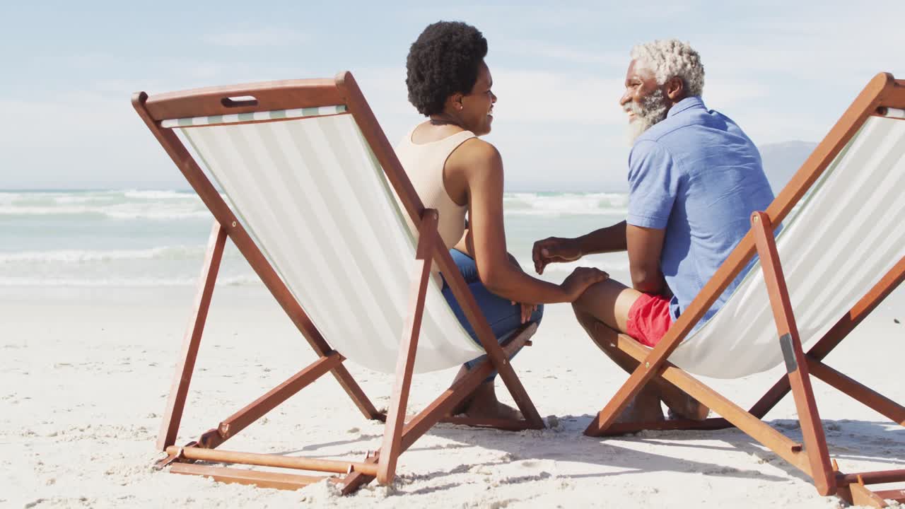 African american couple holding hands and lying on sunbeds on sunny beach