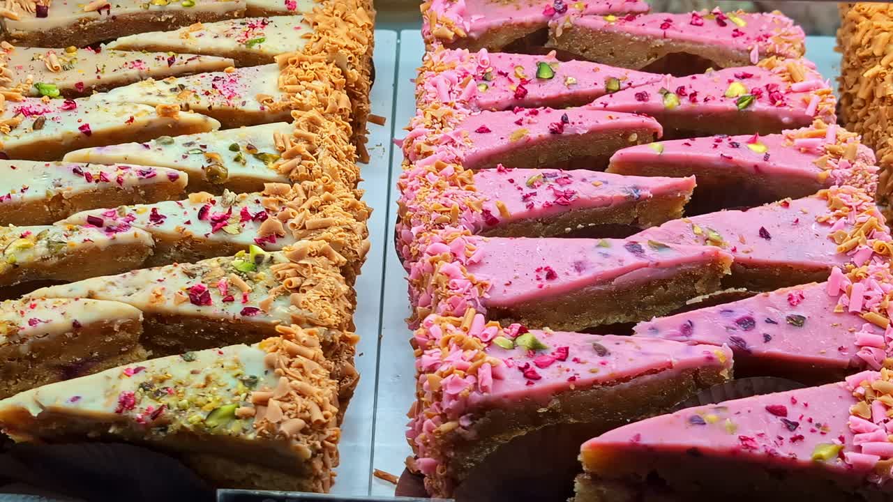 Una panorámica lenta revela una vibrante selección de pasteles variados con glaseado rosa y blanco, aderezos rallados y frutos secos en un expositor de panadería.