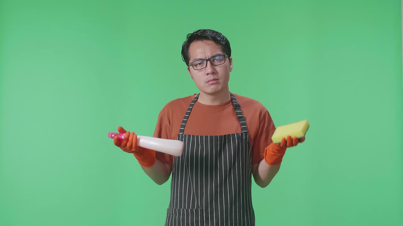 초록색 스크린 스튜디오에 서 있는 스프레이와 부 스폰지를 보여주는 앞옷을 입은 혼란스러운 아시아 남성 집주인