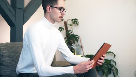 joven con bigote en suéter beige sentado en un sofá en la sala de estar y usando una tableta o un dispositivo inteligente en una habitación moderna y luminosa