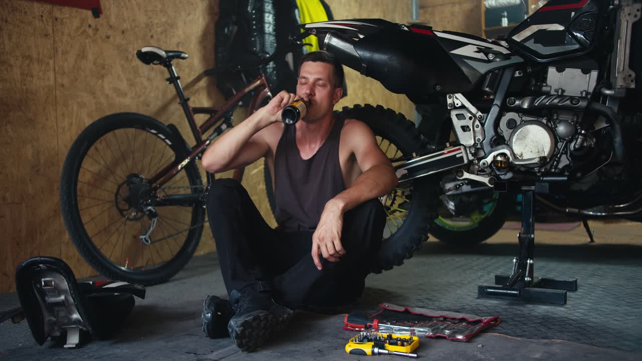 A male mechanic with a short haircut in a gray T-shirt drinks a drink from a dark glass bottle while sitting near his motorcycle in his workshop. Rest during a break during motorcycle repair and maintenance