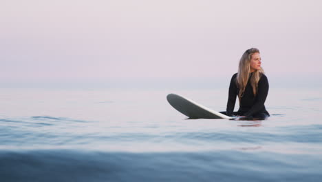 Mujer-Vestida-Con-Traje-De-Neopreno-Sentada-Y-Flotando-Sobre-Una-Tabla-De-Surf-En-El-Mar-Mientras-Las-Olas-Rompen-A-Su-Alrededor.