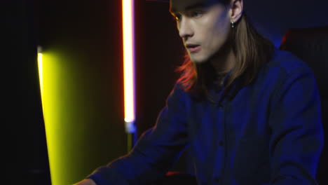 close up view of young serious man wearing cap and playing a game on the computer in a room with colorful neon lamps on the wall 1