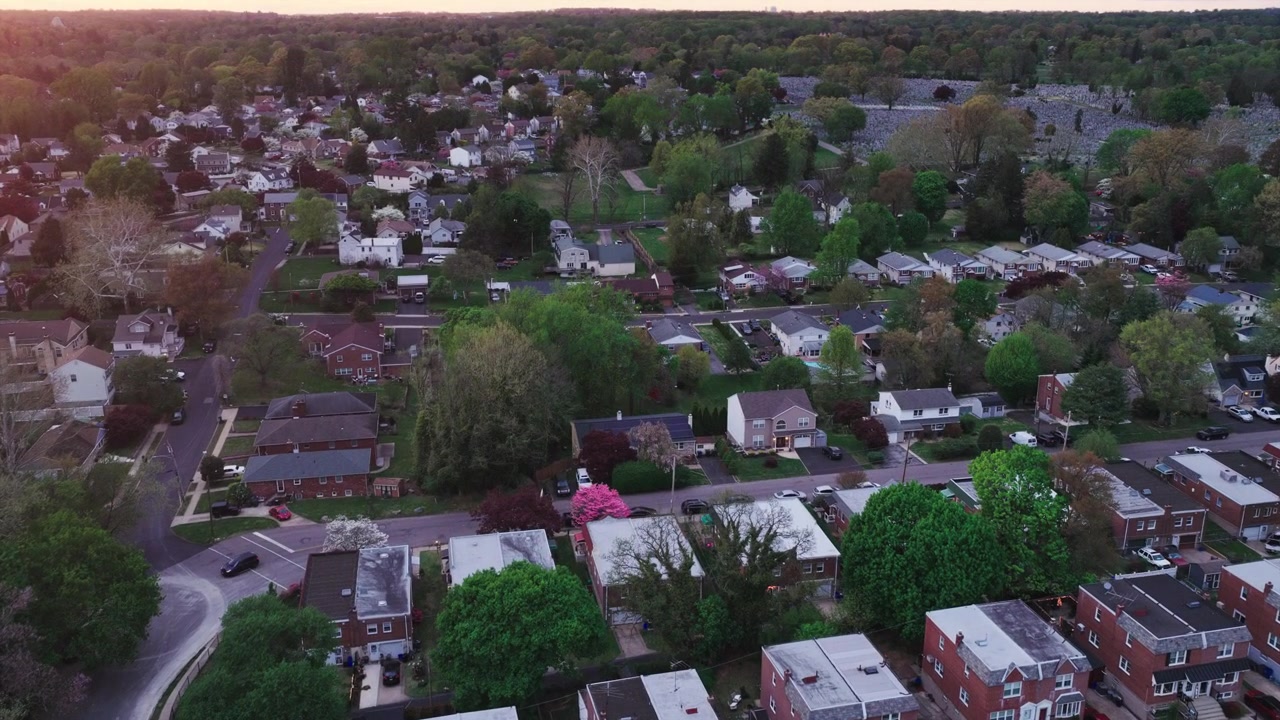 Vista aérea de drone ao pôr do sol de um bairro suburbano da Filadélfia, apresentando casas geminadas de tijolos, ruas arborizadas, jardins e folhagem exuberante de primavera na hora dourada.