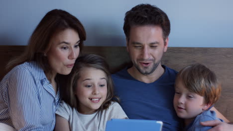 Vista-De-Cerca-De-Los-Padres-Caucásicos-Sentados-En-La-Cama-Con-Su-Pequeño-Y-Lindo-Hijo-E-Hija,-Viendo-Algo-En-La-Tableta-Y-Hablando-Por-La-Noche