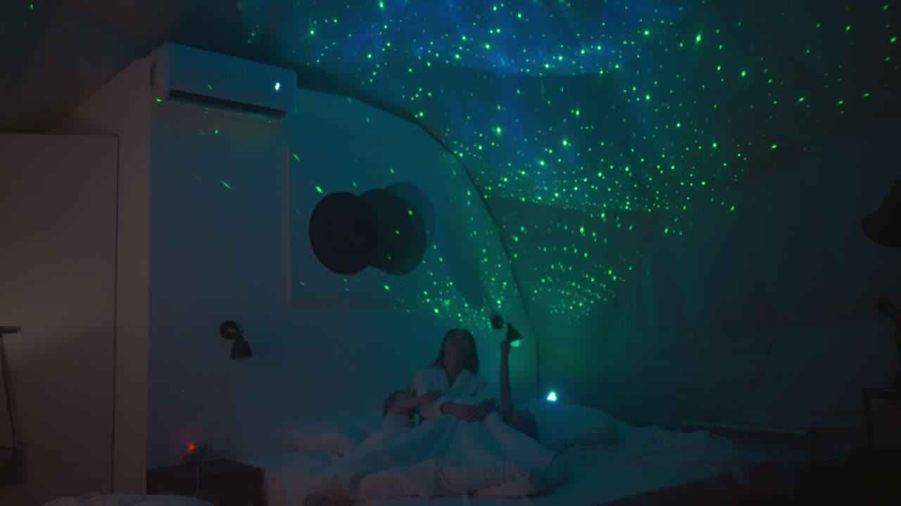 작은 아이들과 함께 편안한 침대에 누워있는 여성은 벽에 비머가 표시하는 별을보고 있습니다. 아이들은 가운 조명으로 방에서 침대에 준비합니다.