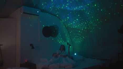 mujer se relaja en una cama cómoda con niños pequeños mirando las estrellas mostradas por el radiador en la pared. los niños se preparan para la cama en una habitación con iluminación apagada