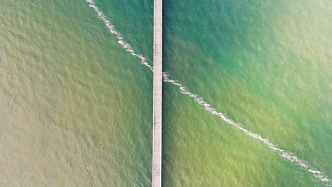 Birds-eye-view-flyover-of-a-long,-wooden-pier-stretching-into-the-gorgeous-clash-of-denim-blue-and-lime-green-colours-mixing-in-the-vast-ocean-on-a-beautifully-sunny-day