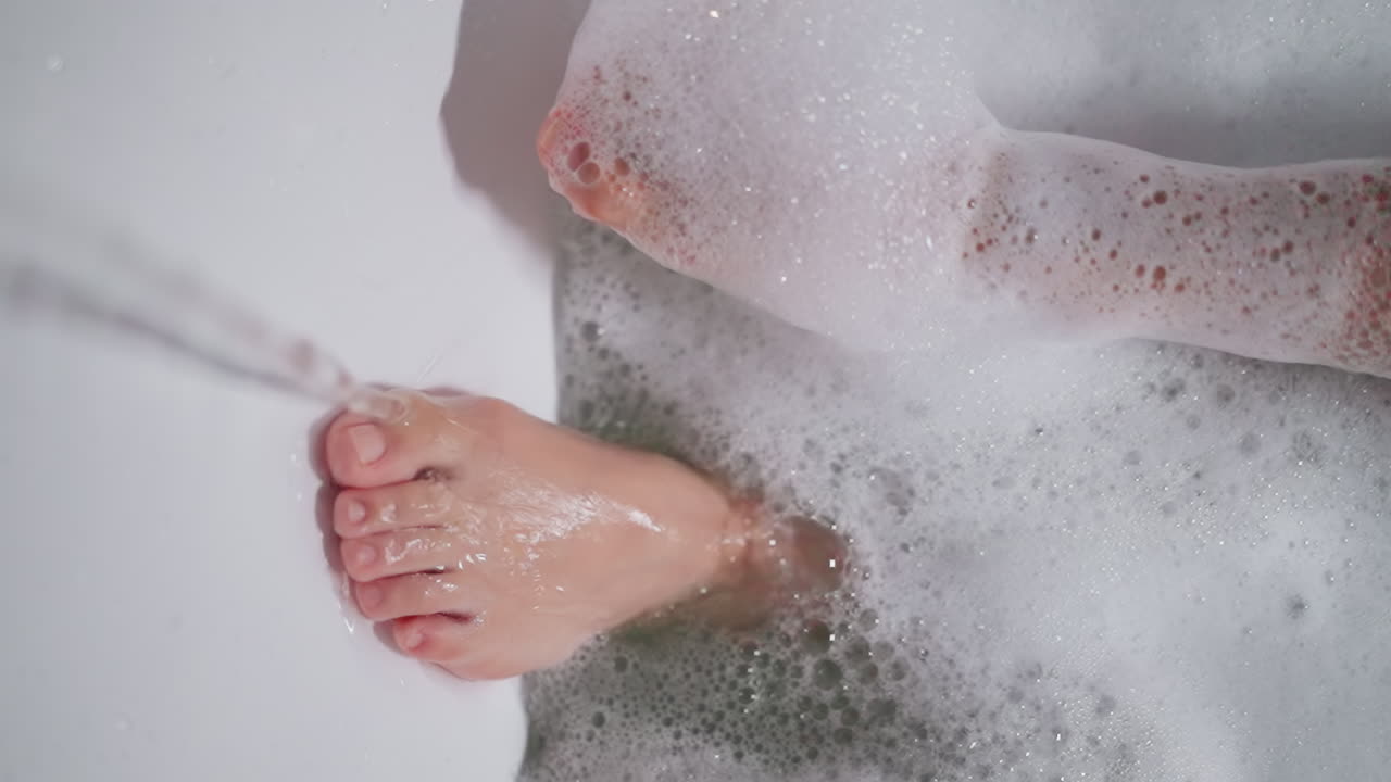 Woman feet in warm foamy bathtub water closeup. Lady enjoys body care procedures washing legs in cozy bathroom closeup. Pleasure of hygienic routine