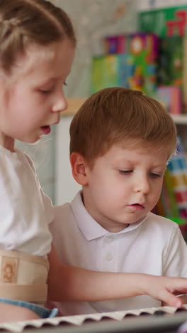 toddler kid sighs while elder sister shows boring musical chords in children room. demotivated little boy looks at girl with corsage playing music