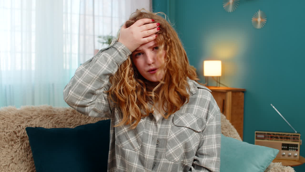 Woman sits on sofa making facepalm gesture feeling disappointed frustrated after unexpected failure