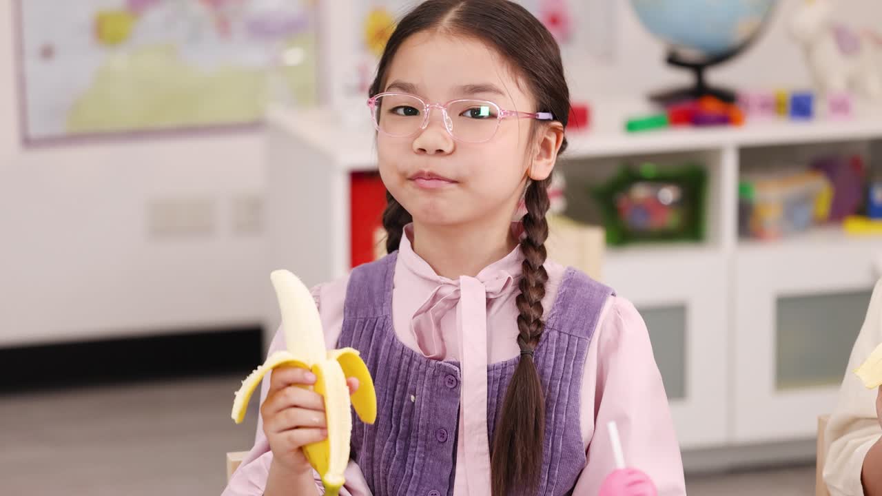 Asian schoolgirl with glasses peels and eats banana in brightly lit classroom, mid-shot, natural light