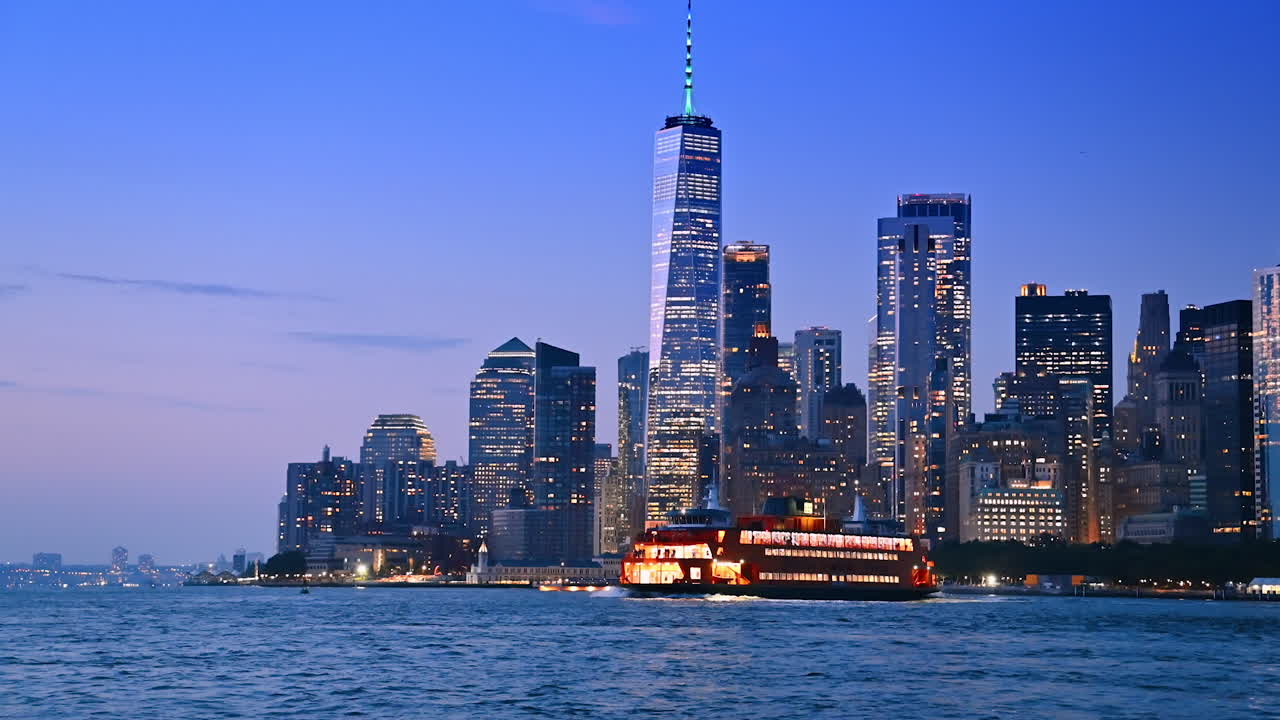 Multi-deck riverboat with switched-on light moves by the East River at dusk. Stunning skyline of luminous Manhattan at backdrop. New York, USA