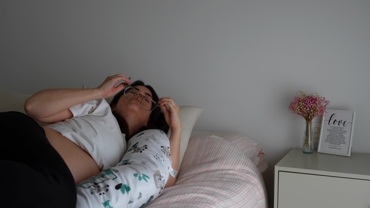 Pregnant woman stretches in bed with glasses as morning light enters cozy bedroom