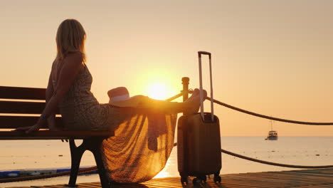 a romantic woman in a light dress with a burly bag sits on the dock looks at the sunrise over the se