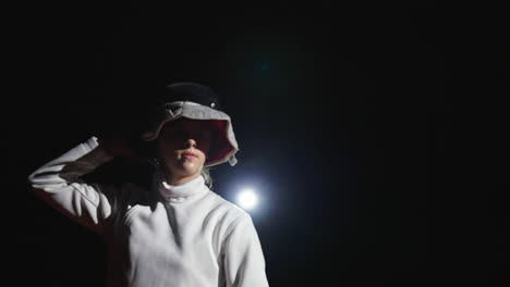 fencer in a black mask and white uniform, standing in front of a bright light