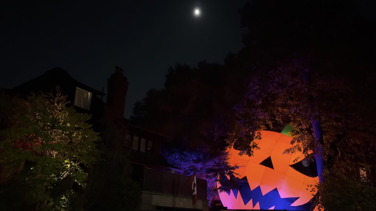 Fixed night shot in a Toronto street showing a huge inflatable pumpkin Halloween decoration under the moonlight, with trees and houses behind