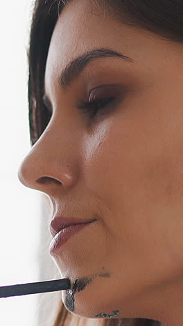 pretty brunette artist with grey paint on hand touches chin with paintbrush looking at drawing on light background in studio extreme closeup slow motion