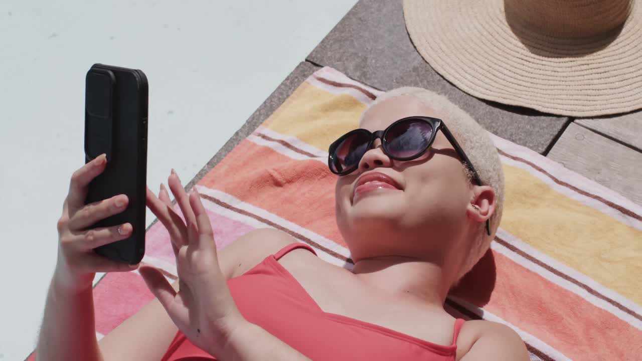 Happy biracial woman with sunglasses using smartphone and sunbathing on towel in slow motion