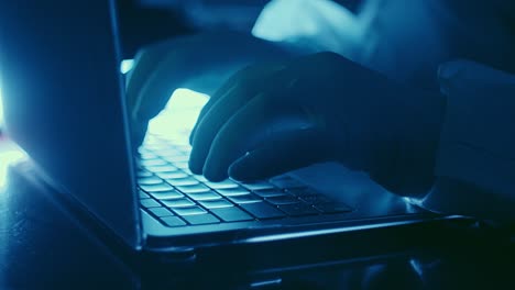 hacker typing laptop keyboard in protective gloves and suit, blue light closeup