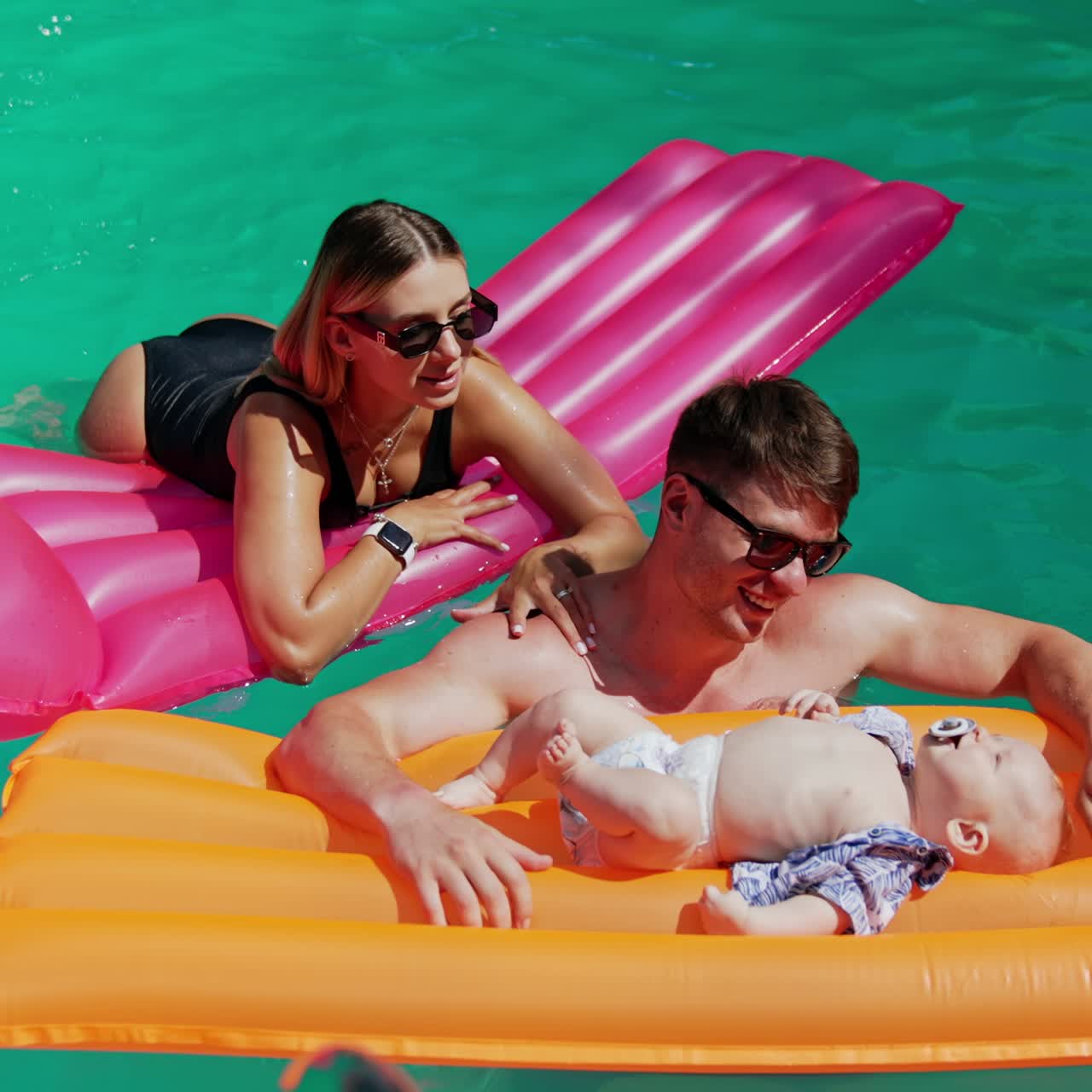 Caucasian parents spending time in the pool with their baby. Infant lies on the mattress waving feet and hands