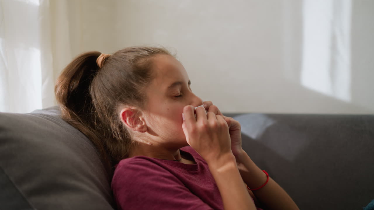 Side view of little girl sneezing into tissue paper while lying on couch, resting at home, natural light filters through window, showing symptoms of illness like cold or flu