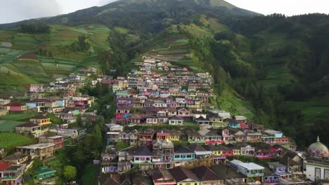 Aerial-backwards-flight-showing-colorful-Nepal-Van-Java-Tourist-Village,-Magelang,-Indonesia---Green-Mountain-with-plantation-surrounding-indonesian-City