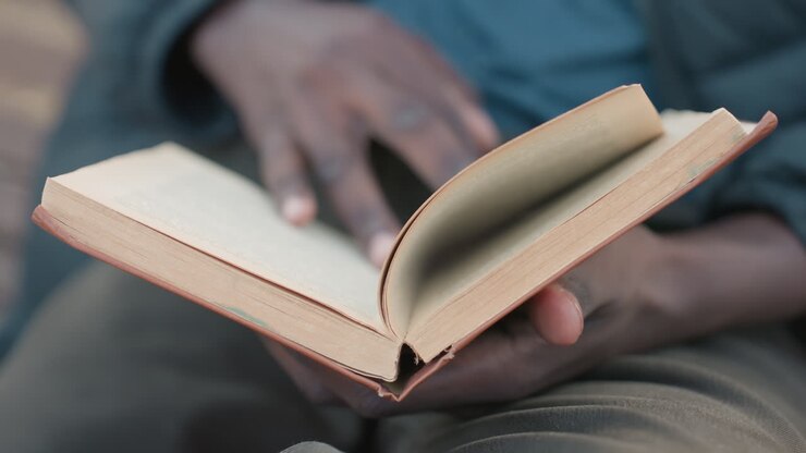 Close up of hands holding open novel while reading outdoors, pages slightly worn, blurred view of reader in background wearing casual clothing, relaxed atmosphere