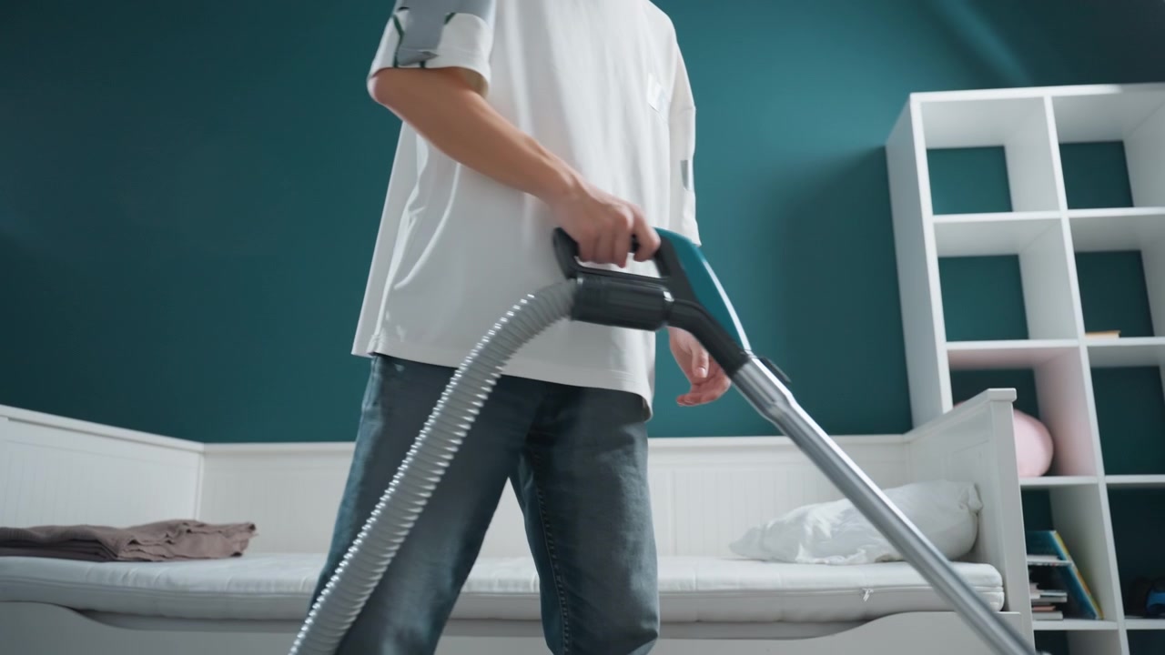 Asian young male vacuuming carpeted floor, energetic movement with long strokes over rug near bed, teal wall and storage shelf, focused domestic chore capturing motion of hose and nozzle