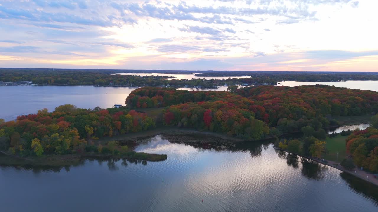 Lake Minnetonka is framed by a vivid tapestry of fall foliage, with rich crimson, amber, and golden trees reflecting the beauty of autumn from above