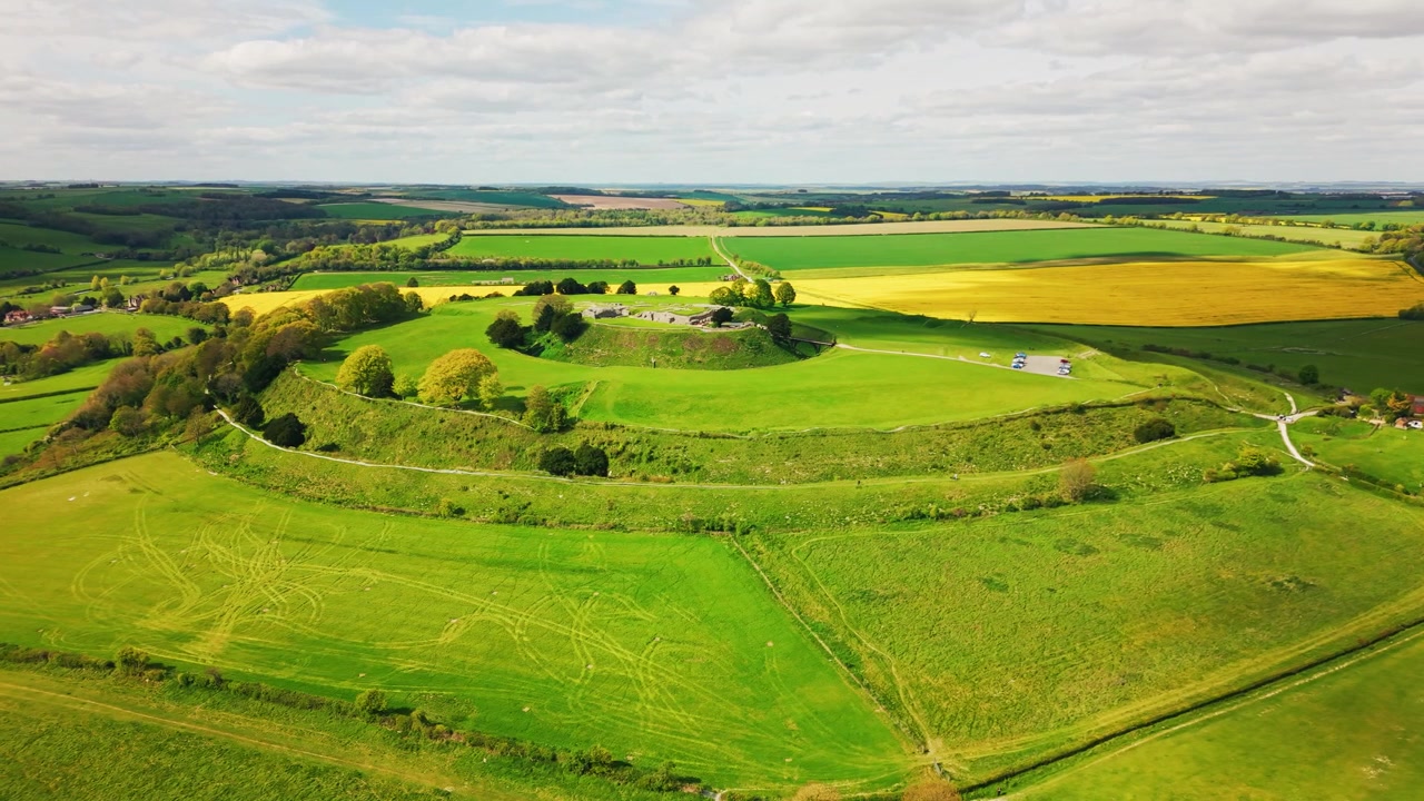 Wide aerial drone shot of Old Sarum near Salisbury in Wiltshire, showing the ancient hillfort earthworks, historic ruins, green grassland, farmland, yellow crop fields and surrounding English countrys