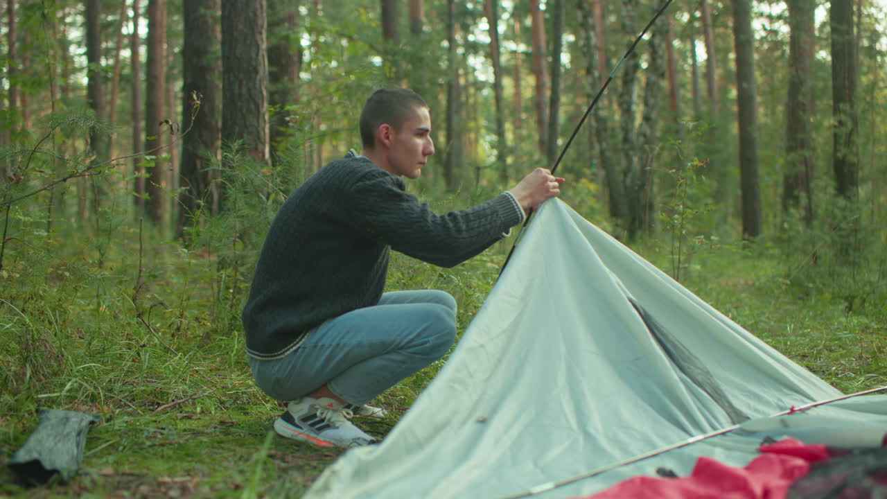 Young student crouches attentively in forest, gripping flexible pole while fixing tent fabric with concentration, scene captures focused outdoor activity surrounded by trees