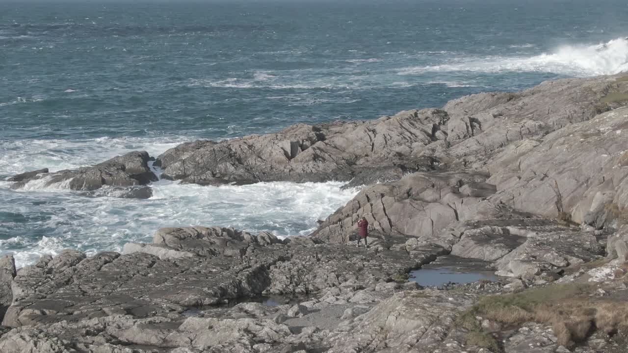 Aerial fly by of a person standing on the rocky shoreline taking pictures while the big waves crash into the coastline during a cold day