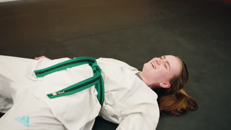 a young female girl combat fighter taekwondo karate martial arts student struggles to get up after long exhausting physical training smiles and laughs as the camera moves out