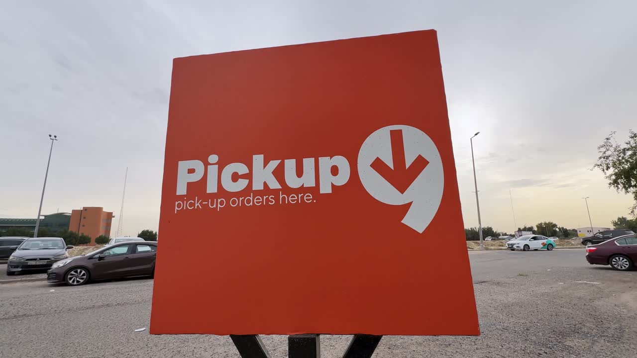 Wide low-angle shot of a bold orange pickup orders sign on a metal stand in an open urban parking lot under an overcast sky, with parked cars and buildings in the background